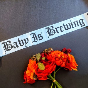 May include: A light blue satin sash with black text that reads "A Baby Is Brewing" and a black silhouette of a baby's footprints in a circle.