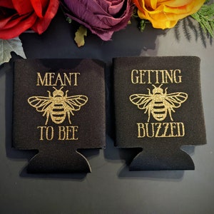 May include: Two black foam can coolers with gold glitter designs. One cooler says "Meant to Bee" with a bee illustration. The other cooler says "Getting Buzzed" with a bee illustration.
