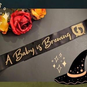 May include: A black satin sash with gold glitter lettering that reads "A Baby is Brewing". The sash has a gold glitter cauldron with two footprints inside.