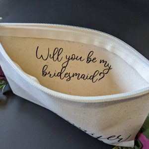 May include: A white canvas zippered pouch with the text "Will you be my bridesmaid?" printed inside. The pouch is open and the zipper is visible.