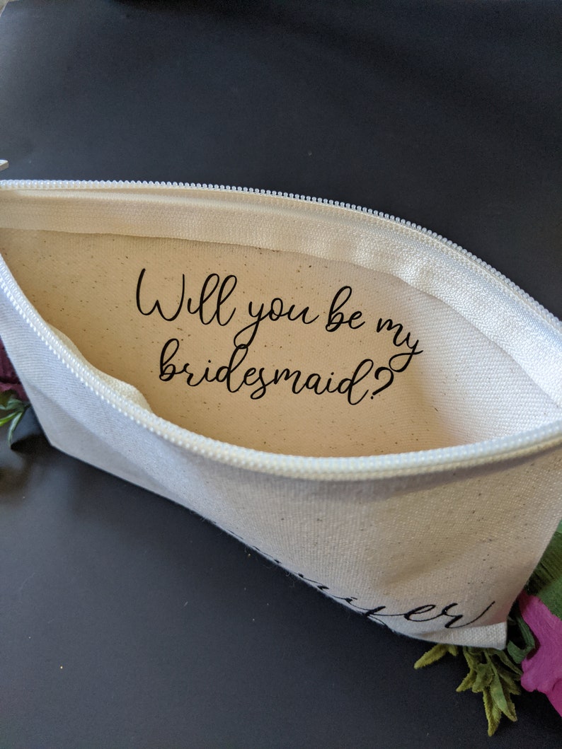 May include: A white canvas zippered pouch with the text "Will you be my bridesmaid?" printed inside. The pouch is open and the zipper is visible.