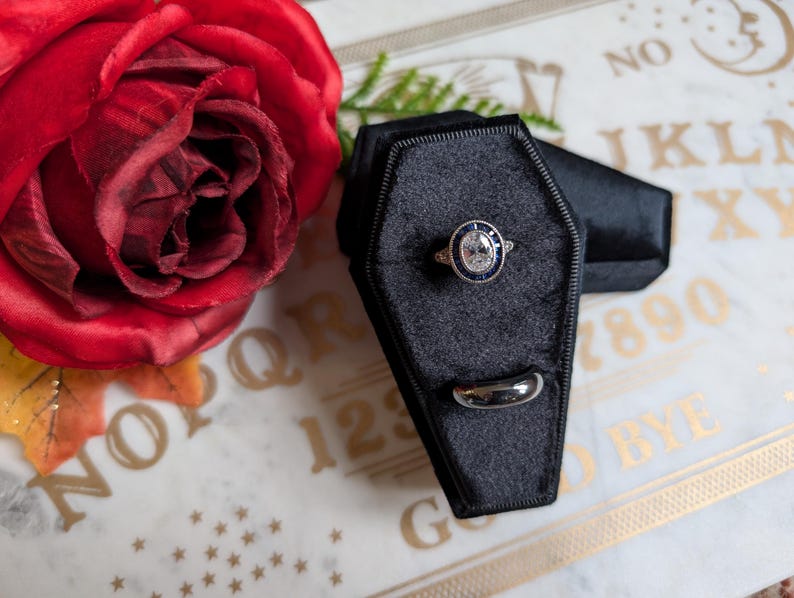 May include: Two black velvet ring boxes shaped like coffins. One box contains a silver ring with a large oval-shaped center stone and smaller stones around the center stone. The other box contains a plain silver band. The boxes are sitting on a white surface with gold lettering that says "Goodbye" and "1234567890".