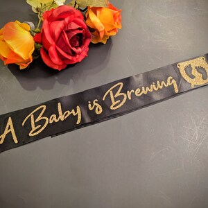 May include: A black satin sash with gold glitter lettering that reads "A Baby is Brewing". The sash has a gold glitter footprint design on the right side.