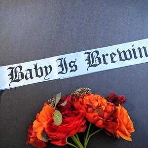 May include: A light blue ribbon with the text "A Baby Is Brewing" in black gothic lettering. The ribbon is lying on a dark gray surface with a bouquet of orange and red flowers in the lower right corner.