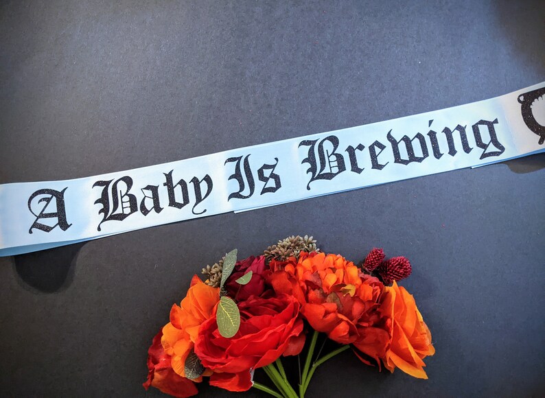 May include: A light blue ribbon with the text "A Baby Is Brewing" in black gothic lettering. The ribbon is lying on a dark gray surface with a bouquet of orange and red flowers in the lower right corner.