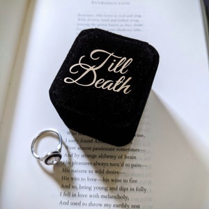 May include: A silver ring with a clear, round gemstone and a black border sits next to a black velvet ring box. The box has the words "Till Death" in elegant script. The items rest on an open book.
