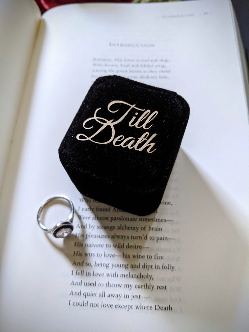 May include: A silver ring with a clear, round gemstone and a black border sits next to a black velvet ring box. The box has the words "Till Death" in elegant script. The items rest on an open book.