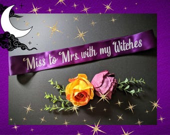 May include: Purple satin sash with the text "Miss to Mrs. with my Witches" in white lettering. The sash is lying on a black surface with two artificial flowers, one yellow and one purple.