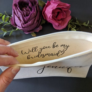 May include: A white canvas zippered pouch with the text "Will you be my bridesmaid?" and the name "Jennifer" written in black cursive. The pouch is open and a hand is holding the zipper pull. Two pink and purple fabric flowers are in the background.