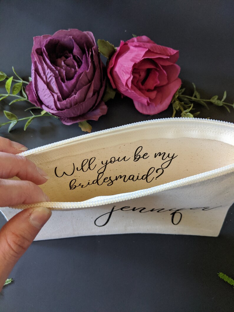 May include: A white canvas zippered pouch with the text "Will you be my bridesmaid?" and the name "Jennifer" written in black cursive. The pouch is open and a hand is holding the zipper pull. Two pink and purple fabric flowers are in the background.