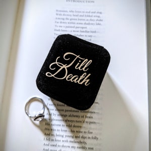 May include: A silver engagement ring rests beside a black velvet ring box with the words "Till Death" in gold script. The ring box sits on an open book with printed text.