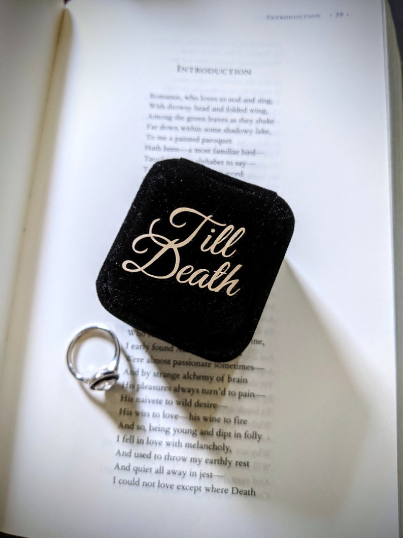 May include: A silver engagement ring rests beside a black velvet ring box with the words "Till Death" in gold script. The ring box sits on an open book with printed text.