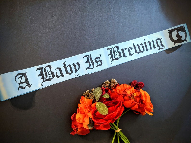 May include: A light blue satin ribbon banner with black text that reads "A Baby Is Brewing". The banner has a small black and white graphic of a baby's footprint at the end.