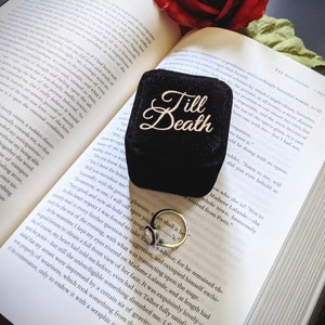 May include: A black velvet ring box with the words "Till Death" in gold script sits open on a book. A gold ring with a white stone rests beside the box. A red rose and greenery are in the background.