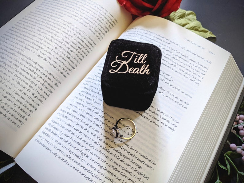 May include: A black velvet ring box with the words "Till Death" in gold script sits open on a book. A gold ring with a white stone rests beside the box. A red rose and greenery are in the background.