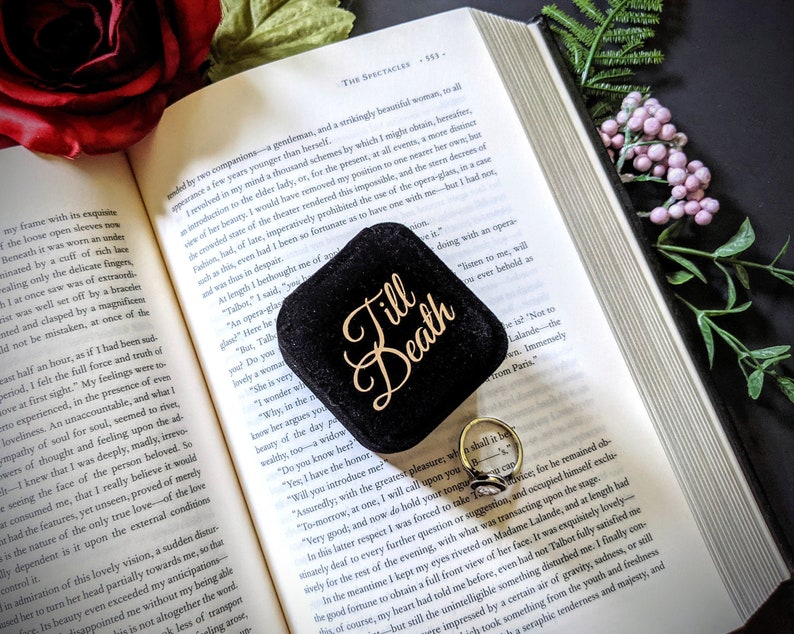 May include: A black velvet ring box with the words "Till Death" in gold script sits open on a book. A gold ring with a black stone is inside the box. A red rose and greenery are in the background.
