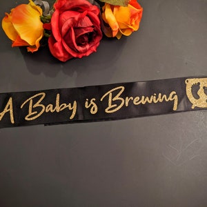 May include: A black satin sash with gold glitter lettering that reads "A Baby is Brewing". A gold glitter cauldron with two baby footprints is on the right side of the sash.