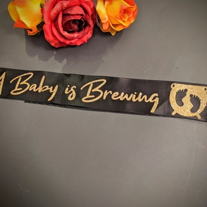 May include: A black satin sash with gold glitter lettering that reads "A Baby is Brewing" and a gold glitter cauldron with two baby footprints inside.