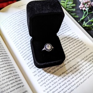 May include: A diamond engagement ring with a halo of blue sapphires sits in a black velvet box, open on a book. The ring's setting is silver, and the book's pages are yellowed.