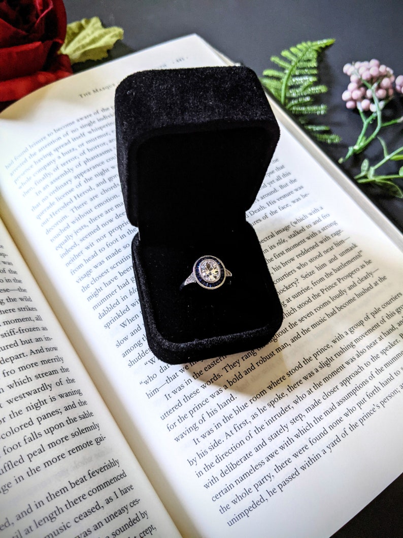 May include: A diamond engagement ring with a halo of blue sapphires sits in a black velvet box, open on a book. The ring's setting is silver, and the book's pages are yellowed.