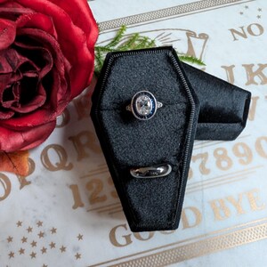 May include: Two black velvet ring boxes shaped like coffins. One box has a silver ring with a blue stone in the center. The boxes are on a white surface with a gold Ouija board.