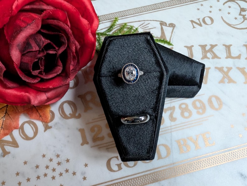 May include: Two black velvet ring boxes shaped like coffins. One box has a silver ring with a blue stone in the center. The boxes are on a white surface with a gold Ouija board.