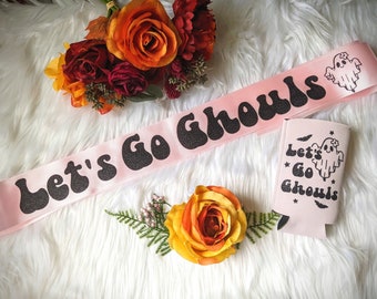 May include: Pink satin ribbon with the text "Let's Go Ghouls" in black glitter letters. A pink can cooler with the same text and a ghost graphic is also pictured.