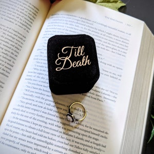 May include: A black velvet ring box with the words "Till Death" in gold script sits open on a book. A gold ring with a white stone is visible inside the box, next to the open book.