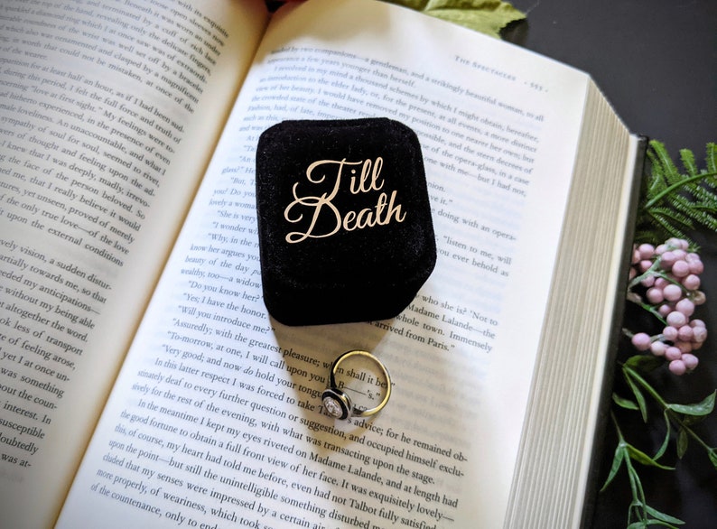 May include: A black velvet ring box with the words "Till Death" in gold script sits open on a book. A gold ring with a white stone is visible inside the box, next to the open book.