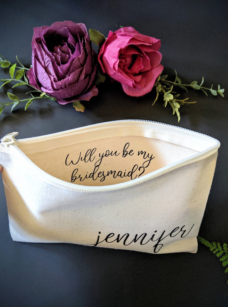 May include: White canvas zippered pouch with the text "Will you be my bridesmaid?" and the name "jennifer" written in black cursive.