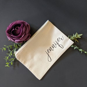 May include: A white canvas zippered pouch with the name "Jennifer" written in black cursive script. The pouch is on a dark gray surface with a purple flower and green leaves.