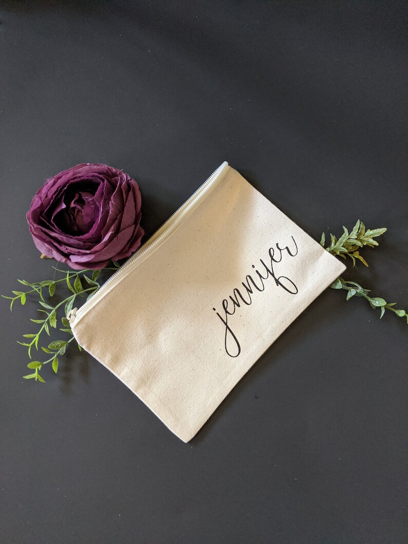May include: A white canvas zippered pouch with the name "Jennifer" written in black cursive script. The pouch is on a dark gray surface with a purple flower and green leaves.