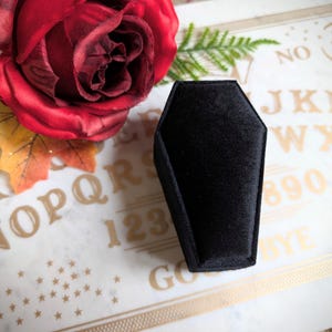 May include: A black velvet coffin-shaped ring box on a white surface with gold lettering that says "NO, J, K, L, M, W, X, Y, 8, 9, 0, Y, E, GO".