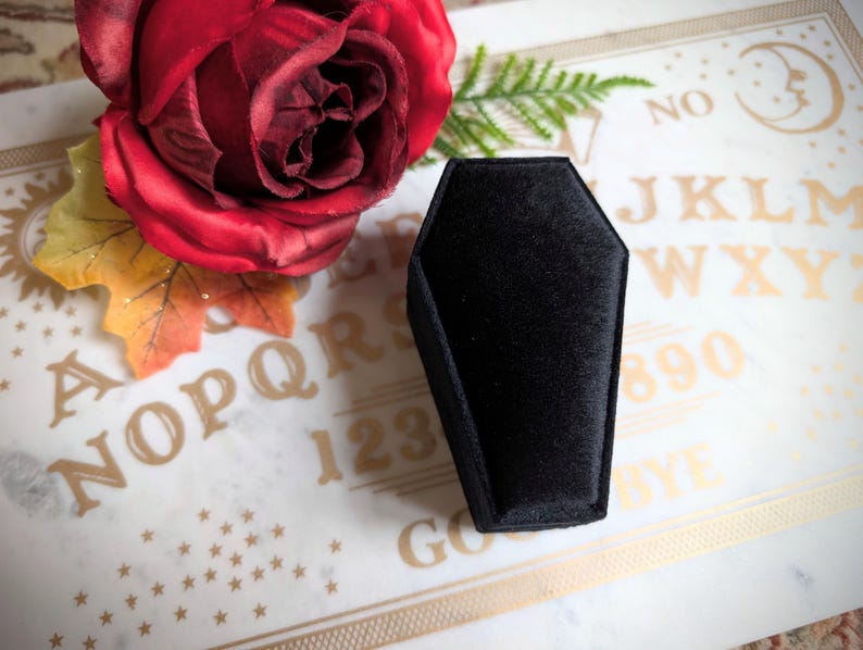 May include: A black velvet coffin-shaped ring box on a white surface with gold lettering that says "NO, J, K, L, M, W, X, Y, 8, 9, 0, Y, E, GO".