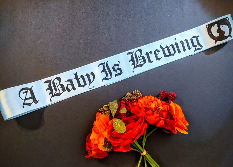 May include: A light blue satin sash with black text that reads "A Baby Is Brewing" and a black silhouette of a baby's footprints in a circle.