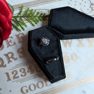 May include: A black velvet coffin-shaped ring box with a silver ring with a large blue stone and a smaller silver band ring inside. The box is sitting on a white surface with gold lettering that says "1234567890 QWERTYUIOPASDFGHJKLZXCVBNM GOOD".