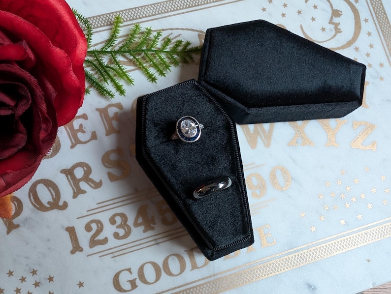 May include: A black velvet coffin-shaped ring box with a silver ring with a large blue stone and a smaller silver band ring inside. The box is sitting on a white surface with gold lettering that says "1234567890 QWERTYUIOPASDFGHJKLZXCVBNM GOOD".
