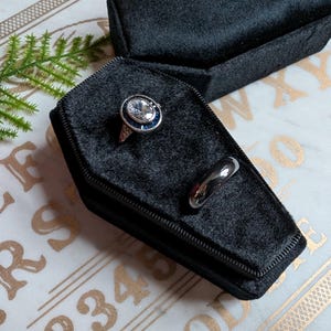 May include: Two rings in a black velvet coffin-shaped box. One ring has a large, clear stone with a blue center. The other ring is a simple silver band.