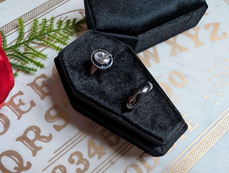 May include: Two rings in a black velvet coffin-shaped box. One ring has a large, clear stone with a blue center. The other ring is a simple silver band.