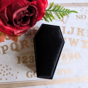 May include: A black velvet coffin-shaped ring box on a white and gold Ouija board with the letters "NO" and "BYE" visible.