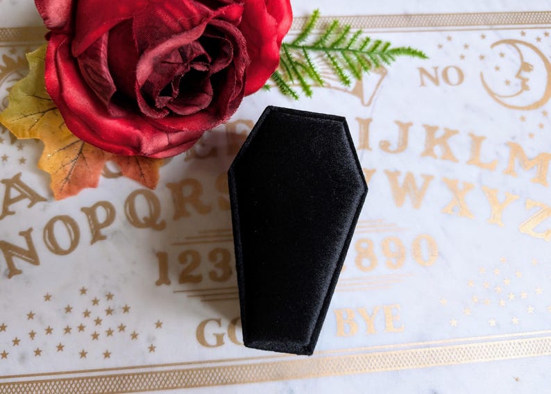 May include: A black velvet coffin-shaped ring box on a white and gold Ouija board with the letters "NO" and "BYE" visible.