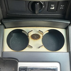 May include: A custom wooden cup holder insert for a car's centre console. It has two circular cup holders and a central, circular area. The wood has a light, natural finish, and the cup holders are dark.