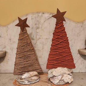 May include: Two decorative Christmas trees. One is wrapped in natural twine, the other in orange twine. Both have a rusty star on top and sit on a base of oyster shells. The trees are on a decorative, crocheted doily.