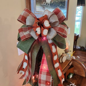 May include: A festive fall-themed tree topper with a large bow made of red and white plaid, green burlap, and brown burlap with a white pumpkin pattern. The bow is tied with a white ribbon and has a white polka dot accent.