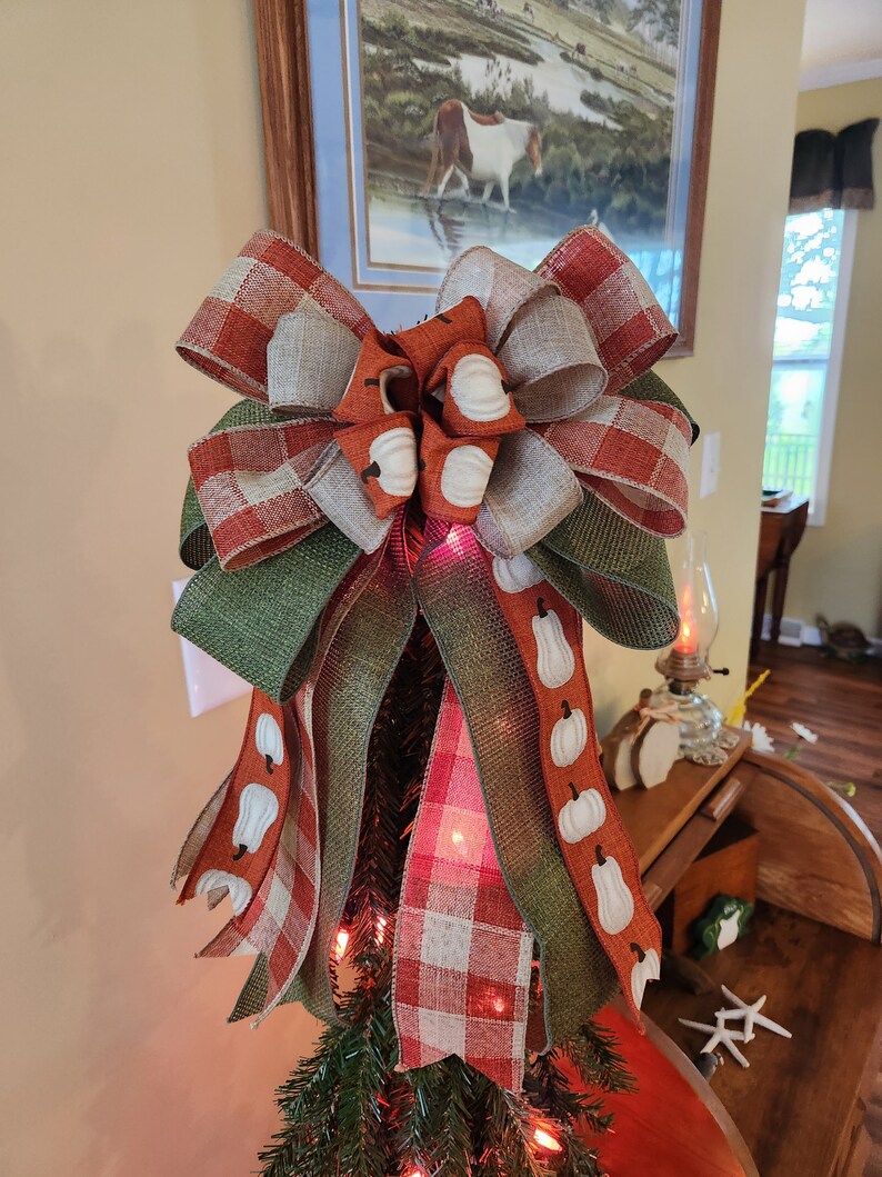 May include: A festive fall-themed tree topper with a large bow made of red and white plaid, green burlap, and brown burlap with a white pumpkin pattern. The bow is tied with a white ribbon and has a white polka dot accent.