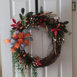 May include: A fall-themed wreath with a natural grapevine base. It features a large orange bow with a black and white checkered pattern, along with red and green leaves, berries, and sprigs. The wreath is hanging on a white door.