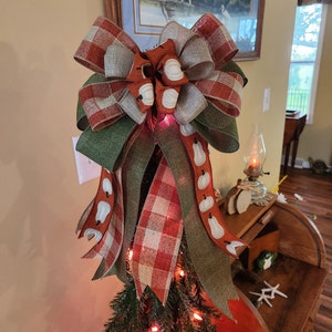 May include: A green, orange, and white plaid bow with a burlap ribbon and white pumpkin accents. The bow is on top of a small artificial Christmas tree with red lights.