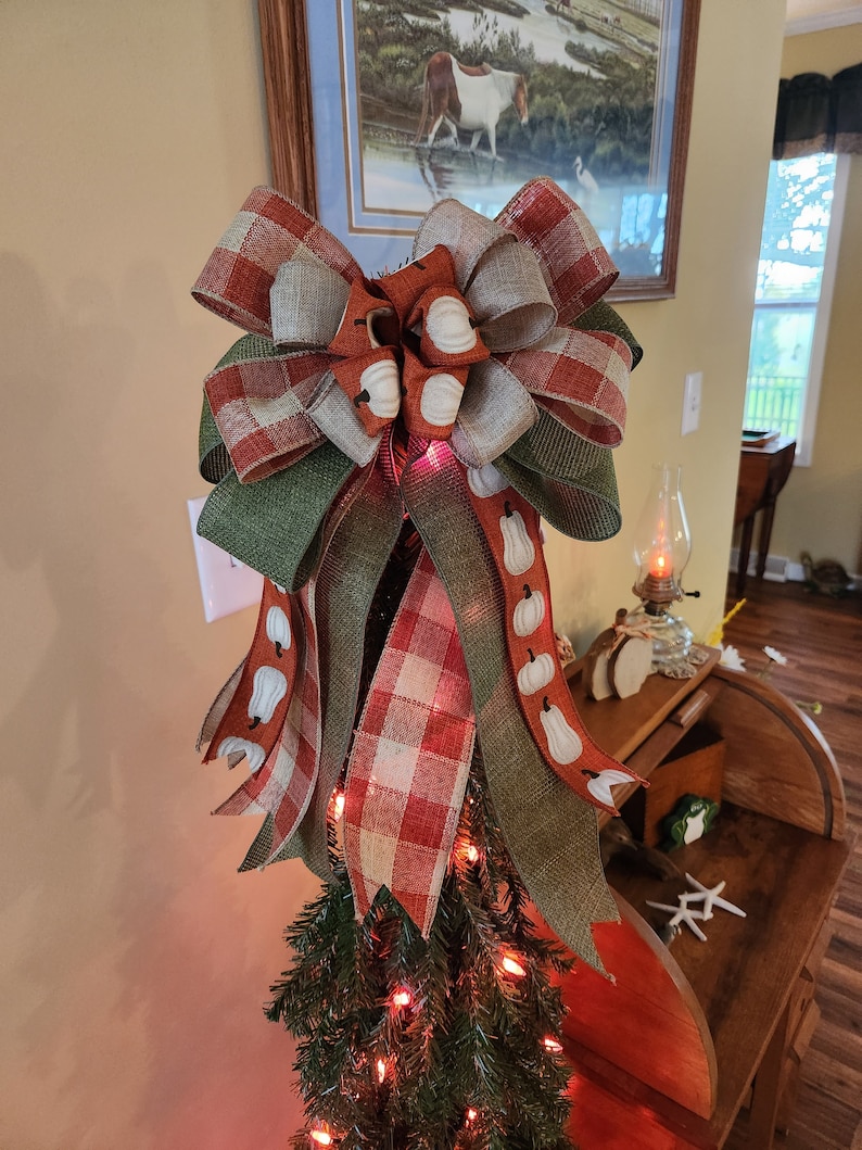 May include: A green, orange, and white plaid bow with a burlap ribbon and white pumpkin accents. The bow is on top of a small artificial Christmas tree with red lights.
