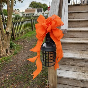 May include: A black lantern with a cage design is decorated with a large orange bow. The lantern is attached to a wooden railing on a set of wooden stairs. A white ghost cutout is on the bottom step.
