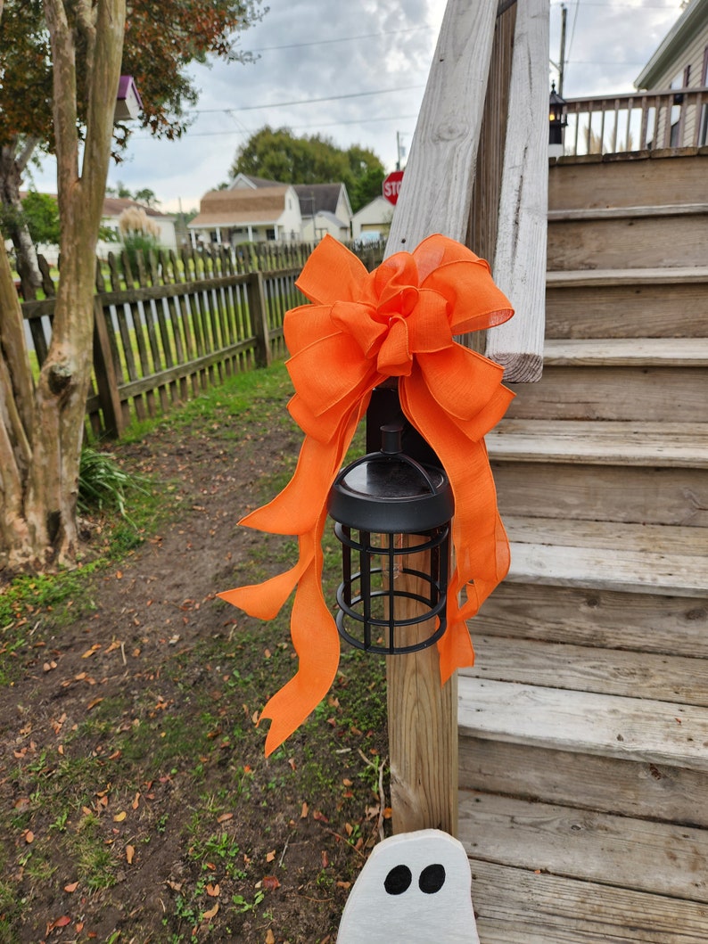 May include: A black lantern with a cage design is decorated with a large orange bow. The lantern is attached to a wooden railing on a set of wooden stairs. A white ghost cutout is on the bottom step.